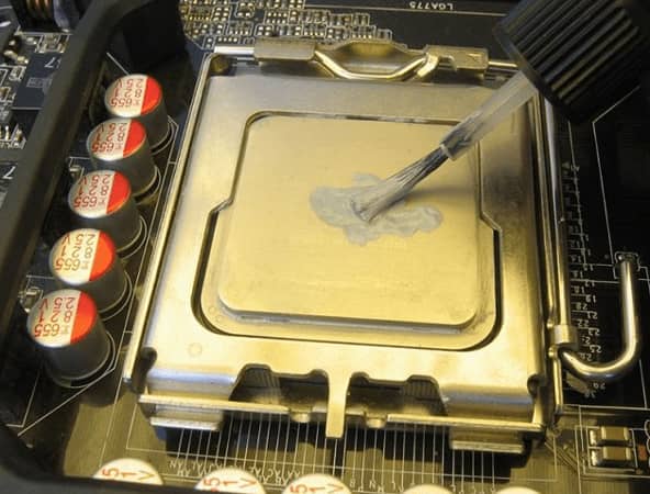 Una mano aplicando pasta térmica sobre un procesador (CPU) que está instalado en la placa base de una computadora, usando una pequeña brocha.