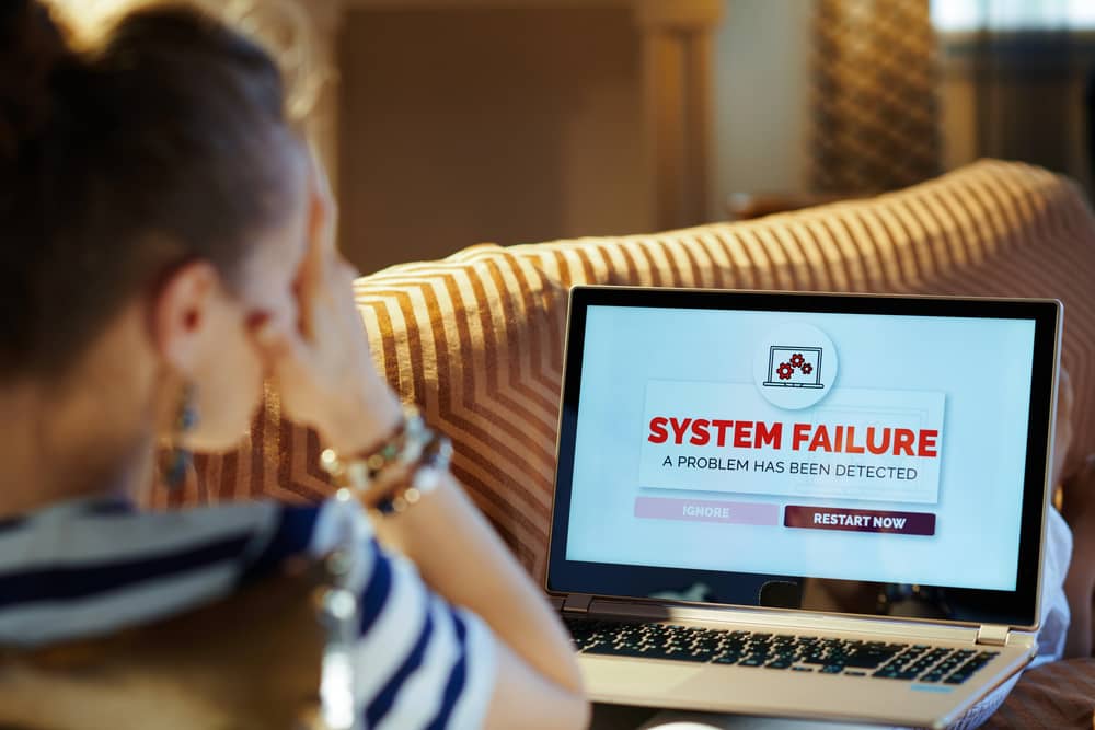 Mi PC se Reinicia Constantemente: ¡Solución Aquí! 9 Una persona sentada en un sofá, con la mano en la frente en señal de preocupación, mirando la pantalla de un portátil que muestra un mensaje de "SYSTEM FAILURE" (Fallo del Sistema) y las opciones de "IGNORE" o "RESTART NOW".