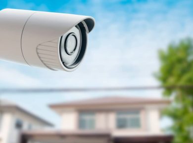 ¿Por qué tener una alarma en el hogar hoy en día ya no es opcional? 11 Cámara de seguridad blanca montada en el exterior, enfocando hacia una casa residencial borrosa en el fondo bajo un cielo azul.